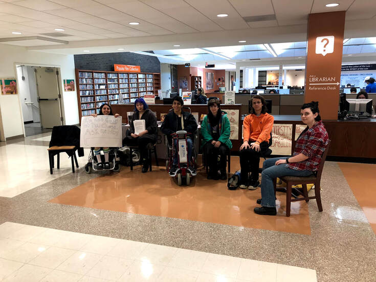     Six Protesters sit in front of the librarian’s desk at the SUNY Purchase Library. Left to right: Sonya is seated in her power chair holding a hand drawn sign reads in bubble letters: “I’m not safe at Purchase, Ask me why.” The text is inside of a speech bubble, a common symbol used in SUNY Purchase branding and logos. Sonya is a petite white femme with shoulder length hair and is holding the sign up which covers her seated body and chin. She is smiling. Next to her is a blue haired white femme holding informational leafletts given to people passing by and interacting with the protest. Next is a medium toned man in a power chair, then a short haired white person in a green jacket, then a white person with shoulder length brown hair in an orange sweatshirt, then finally, sitting at a slight angle is a white woman with short dark hair wearing a red flannel.