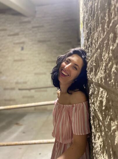 Sonya is standing against a lit stone wall, smiling exuberantly with her profile pivoted slightly to the camera. Her wavy black hair falls to her shoulders and she is wearing a pink & white striped off the shoulder dress. Light washes over her from above, and a railing in blurred in the background.