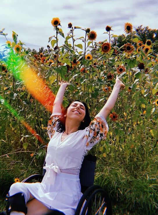 Sonya reclining luxuriously with arms raised to the sky in a black manual wheelchair, in front of tall sunflowers. Her eyes closed, she is smiling with her face tipped up towards the sun. From the left, there are streaks of rainbow light. She is wearing a white blouse with a tie on the front and yellow flowers on billowy sleeves, as well as a white skirt and back leg braces that come up to her knee.