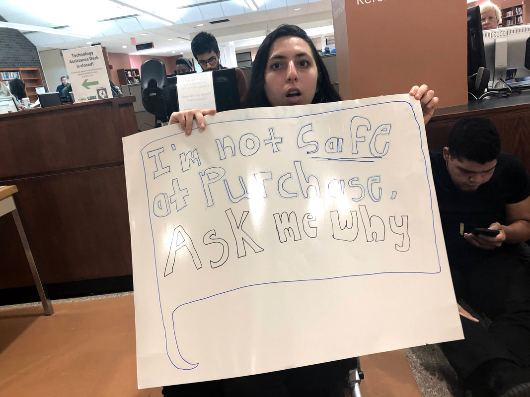  Photograph of Sonya holding a hand drawn sign reads in bubble letters: “I’m not safe at Purchase, Ask me why.” The text is inside of a speech bubble, a common symbol used in SUNY Purchase branding and logos. Sonya is a petite white femme with shoulder length hair and is holding the sign up which covers her seated body and chin. Her mouth is open, possibly in speech, and her eyes are determined. Sonya is sitting in front of the librarian’s desk at the SUNY Purchase library.